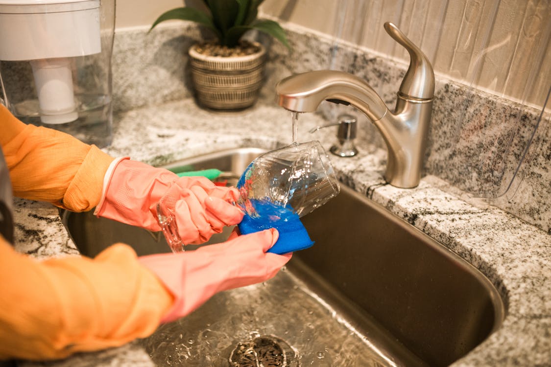 Free Close-up of hands wearing gloves washing a glass in a kitchen sink under a running faucet. Stock Photo