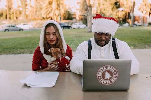 Free A cheerful duo in Santa hats works on a laptop outdoors, embodying holiday spirit. Stock Photo