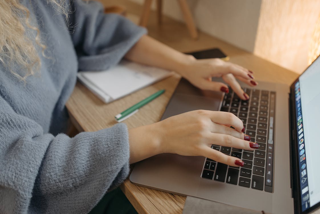 Free Person with red nails typing on a laptop at a wooden desk with a notebook and pencil nearby. Stock Photo