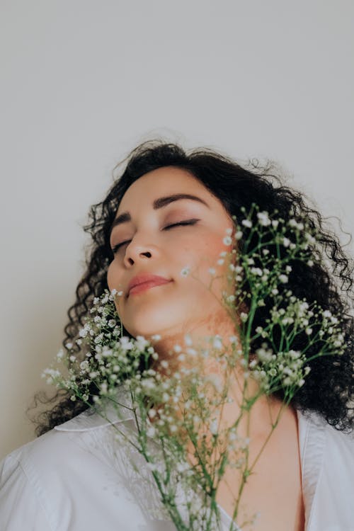 Free Peaceful woman with curly hair posing with delicate flowers, eyes closed in tranquility. Stock Photo