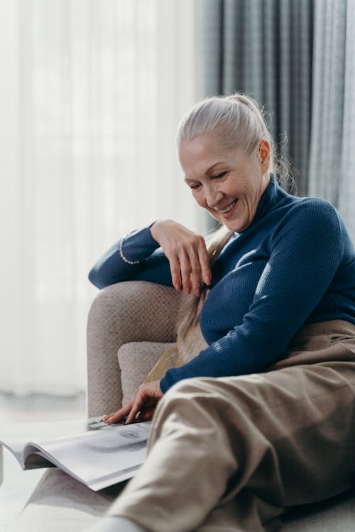 Free Elderly woman enjoying a quiet moment at home, reading a magazine with a smile. Stock Photo