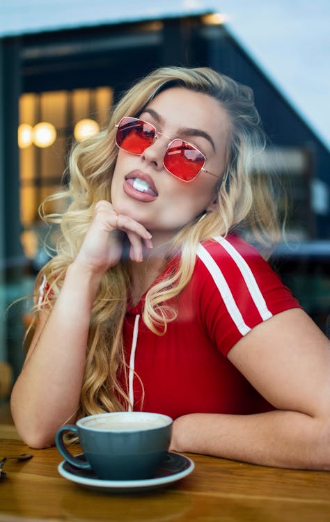 Free A fashionable woman with wavy blonde hair and red sunglasses enjoys her coffee in a cozy cafe. Stock Photo