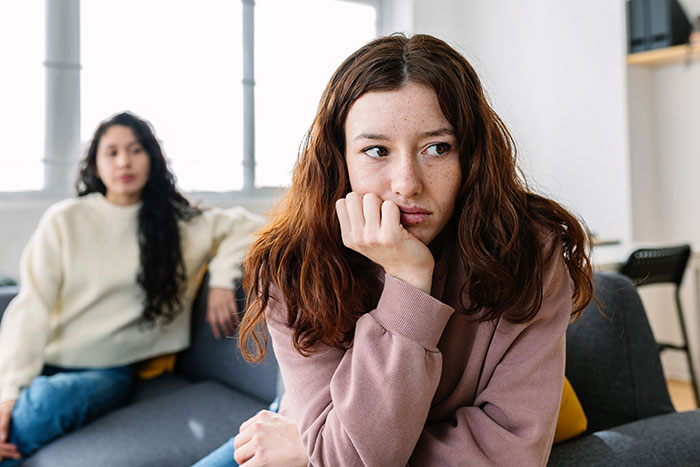 Young woman looking upset and distant on a couch while her sister sits behind her, reflecting a troubled family life.