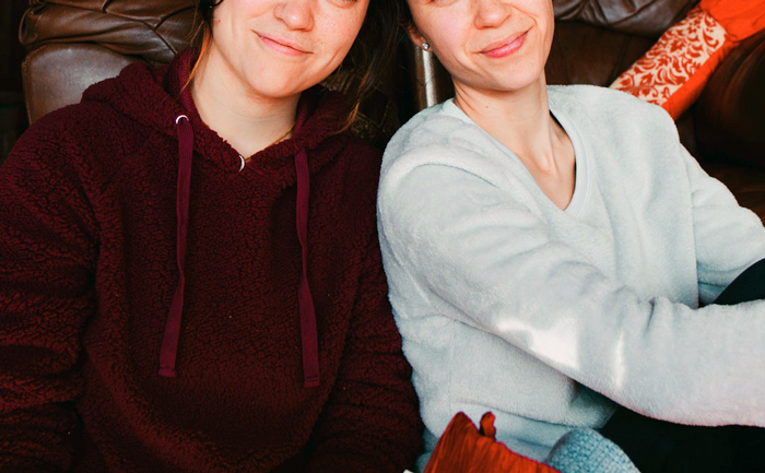 Two sisters sitting closely on a couch, one wearing a maroon hoodie and the other a light gray sweater, smiling gently.