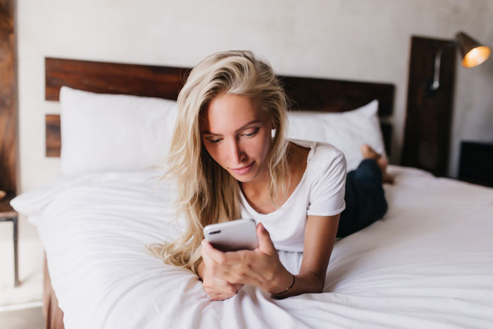 Young woman lying on bed using smartphone, relaxing indoors with a focused expression in a cozy bedroom setting.