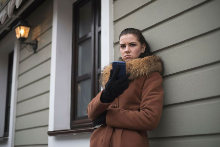 Pregnant woman in a brown coat stands outside house in cold weather, looking upset and holding a smartphone.