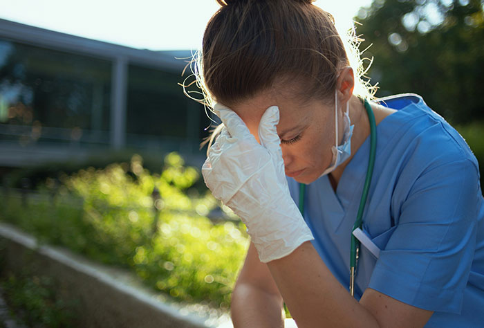 Stressed nurse in blue scrubs with gloves and stethoscope outdoors, reflecting on work nickname causing embarrassment.